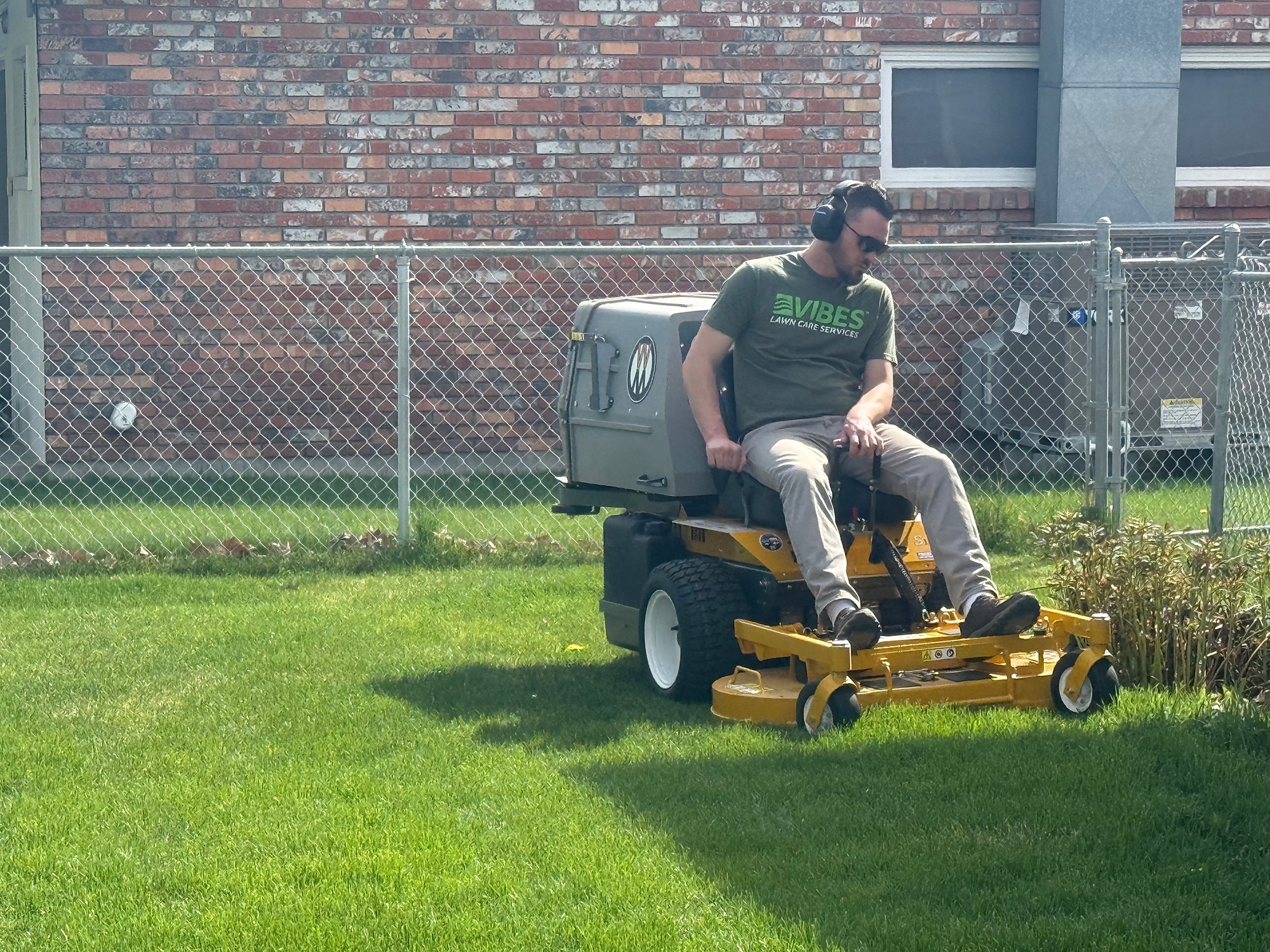 A man on a riding lawn mower trims grass in a residential yard, wearing headphones and a Vibes Lawn Care Services shirt.