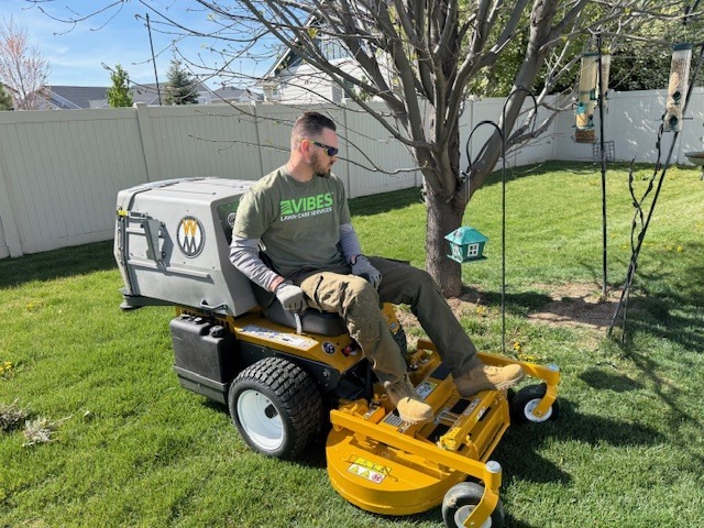 A man sits on a riding lawn mower in a backyard, wearing sunglasses and a green "Vibes" T-shirt, preparing to cut the grass.