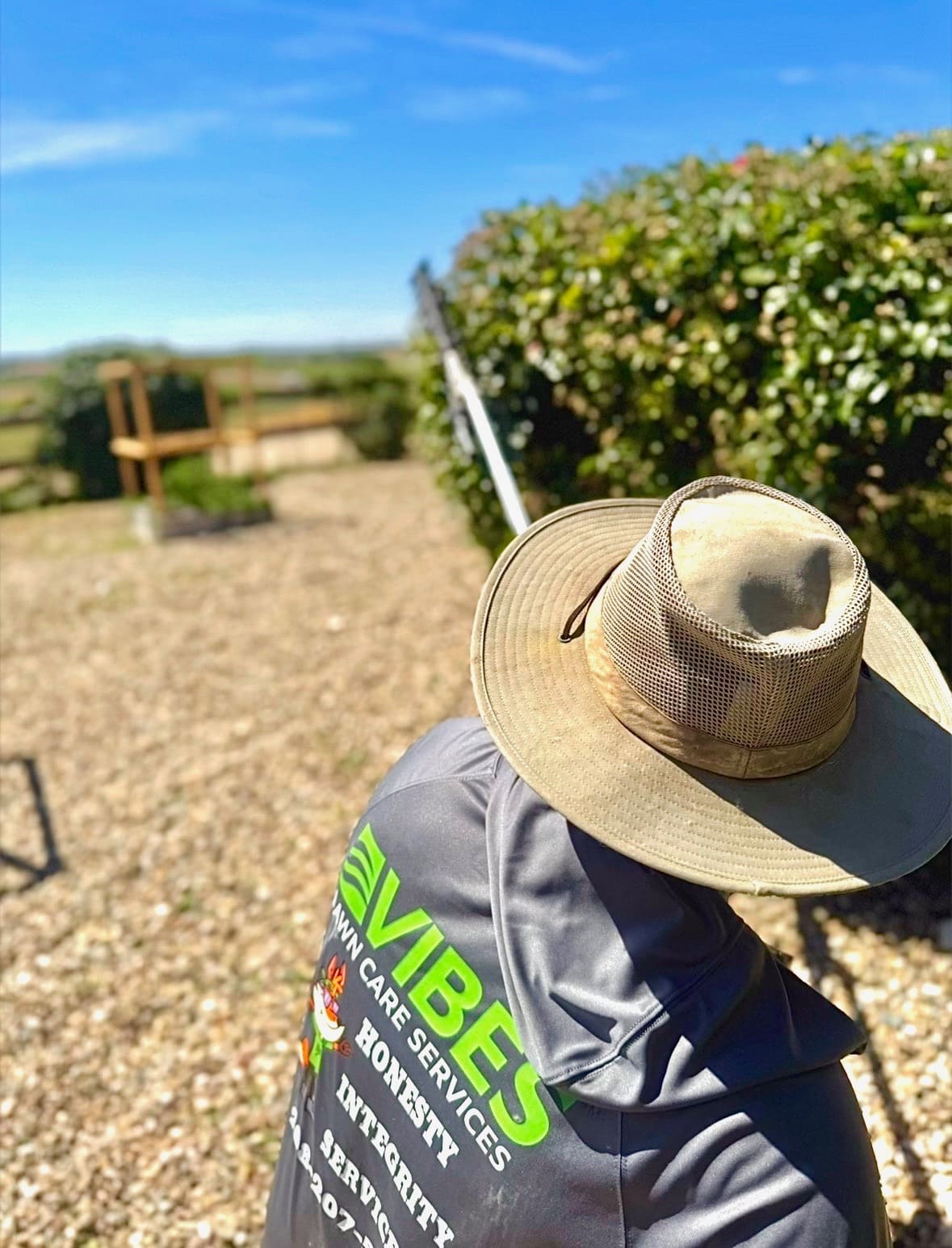 A person in a wide-brimmed hat and a shirt with text "Vibes Lawn Care Services" faces a sunny garden, pruning bushes. The day is bright and clear.