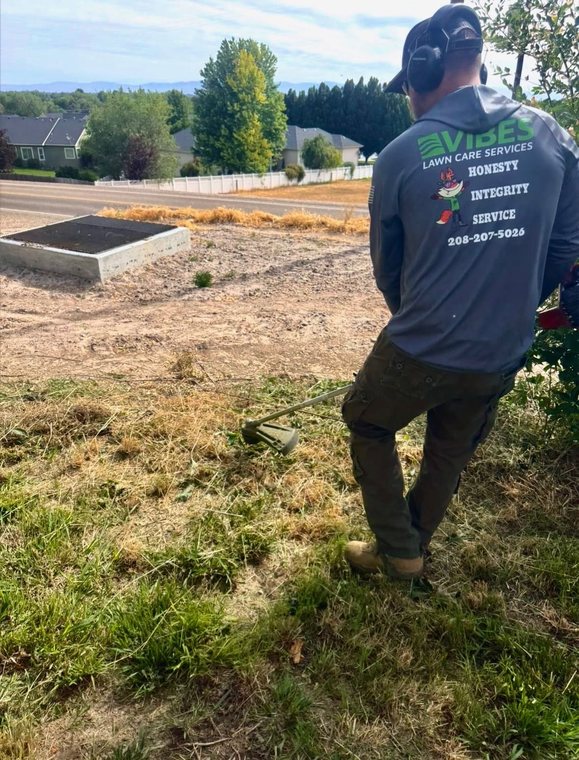 Man in a long-sleeve shirt operates a weed trimmer on a sloped lawn. Background shows a suburban neighborhood with trees and houses.