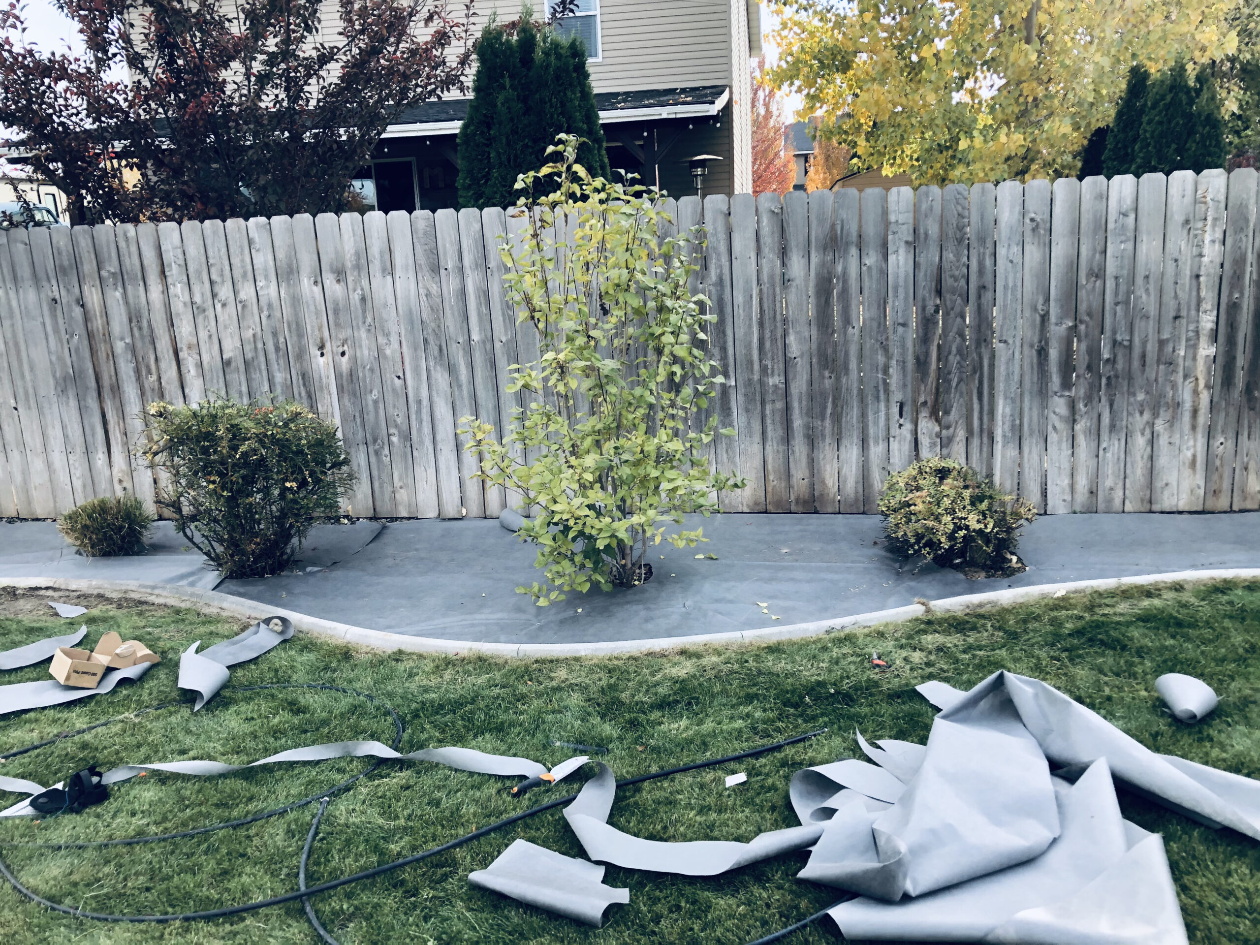 A wooden fence with shrubs and a tree is in the background, while landscaping materials and tools are scattered on a lawn in the foreground.