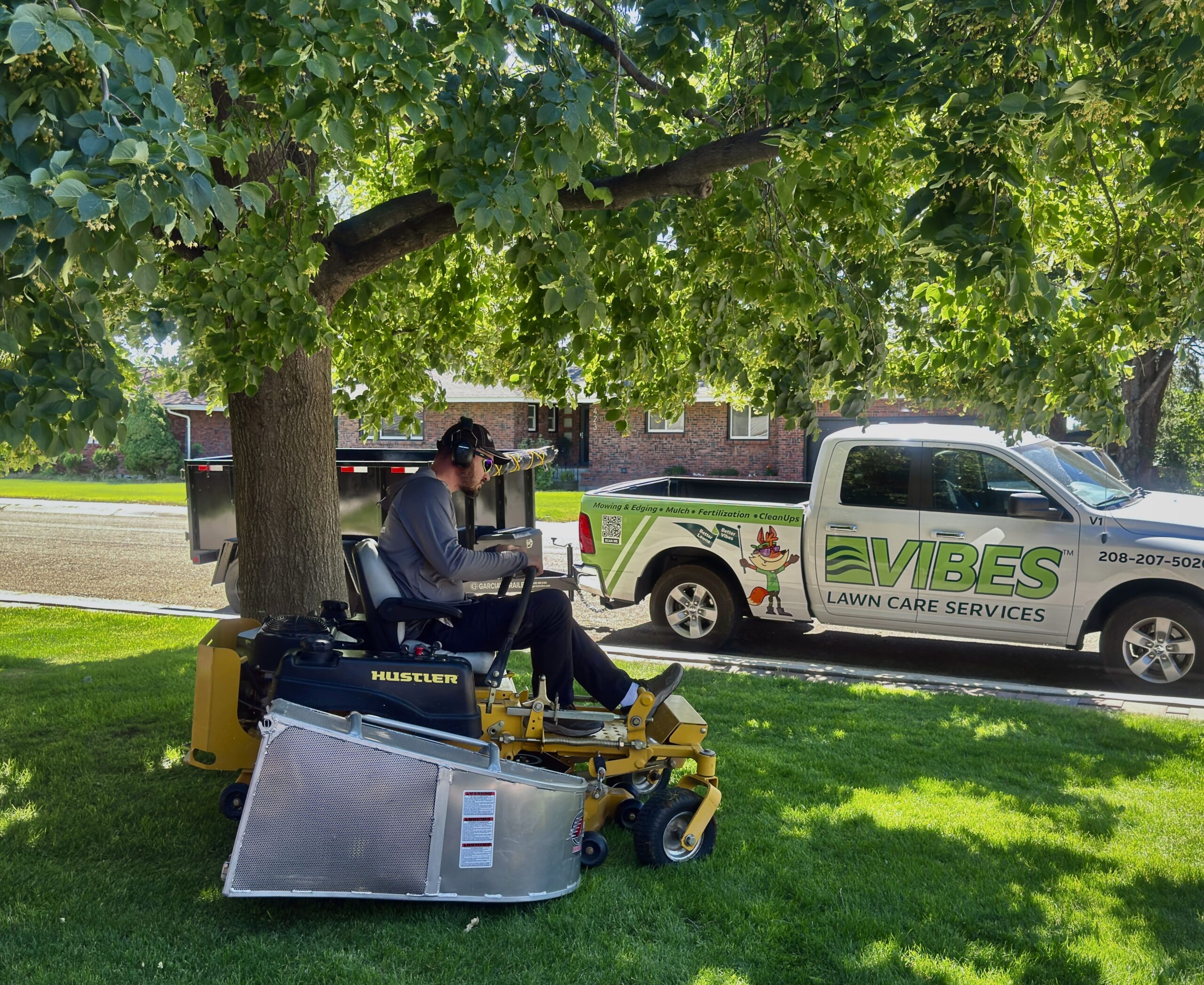 A person operates a ride-on lawnmower beneath a large tree on a sunny day. A truck with "Vibes Lawn Care Services" branding is parked nearby.
