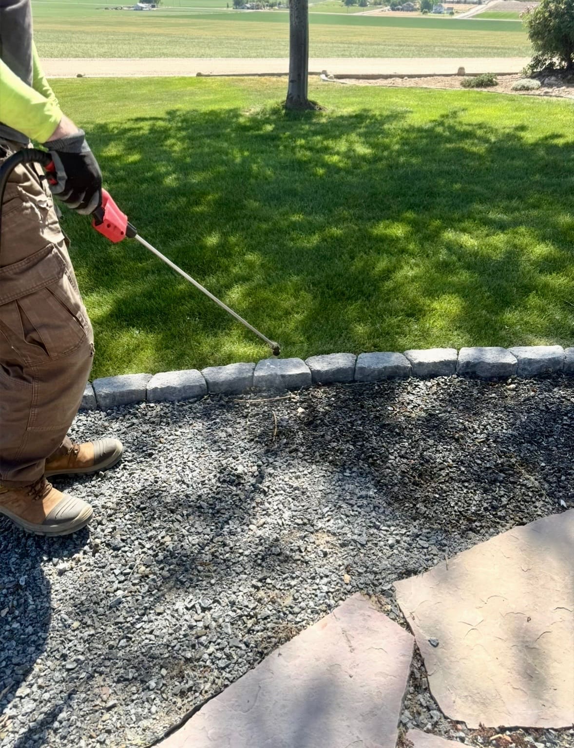 A person in work clothes sprays a gravel path with a hose, adjacent to a garden with green succulents and a stone wall on a sunny day.